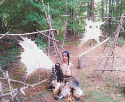 A young Indigenous woman sits on the ground with animal pelts surrounding her and two pelts being stretched behind her