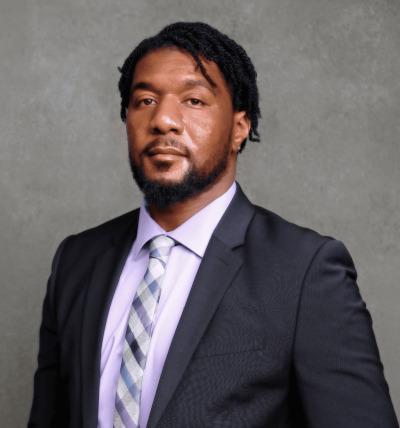 A headshot of a Black man with a beard and mustache wearing a suit with a purple shirt black and white tie