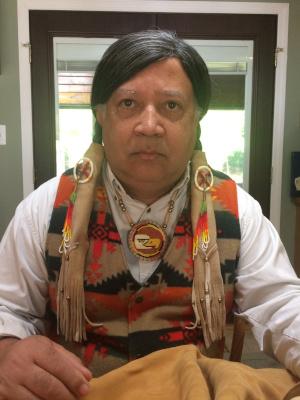 An Indigenous man with long dark hair wearing a colorful vest looks solemnly at the camera
