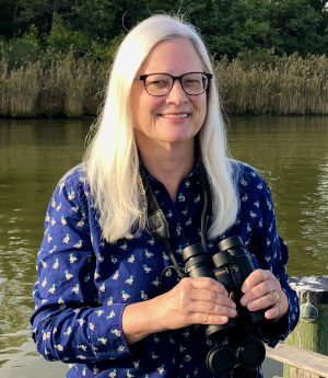 Molly Moore, a woman with long blonde hair and glasses, stands on a pier with water e behind her, wearing a blue shirt and binoculars around her neck