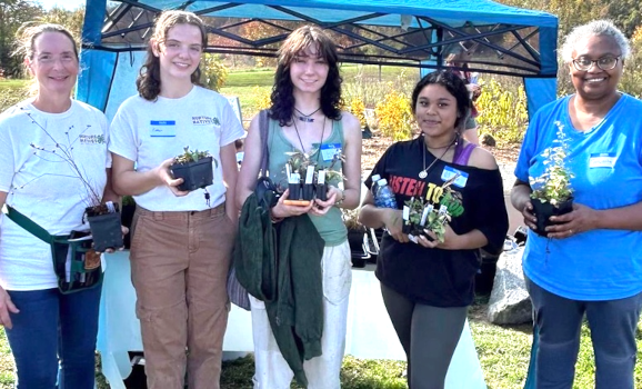 Five women stand outside holding a variety of plants