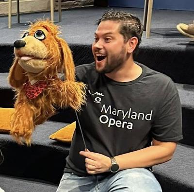 A man in a black Maryland Opera t-shirt holds a large dog puppet
