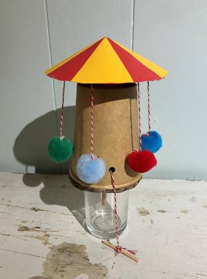 A carousel made from a brown paper cup, with a red and yellow awning, and dangling pom poms