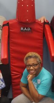 A Black woman with short hair and glasses smiles at the camera while crouching in front of a large red self-defense training dummy.