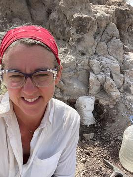 A woman wearing glasses, a red bandana, and a white shirt stands in front of a rocky area