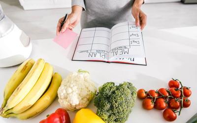 A variety of fruits and vegetables on a counter. A meal planning book is open with a woman standing over it holding a post it note and pen.