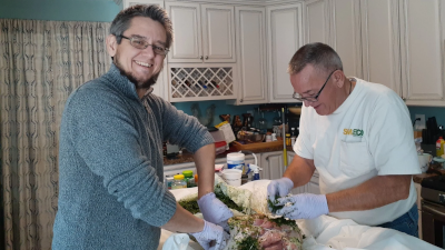Two men stand on either side of a counter, wearing latex gloves and stuffing a ham