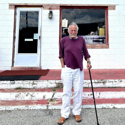 Wayne Karlin, a man with white hair and a beard, stands outside a whitewashed building, wearing a red sweater and white pants holding a walking stick