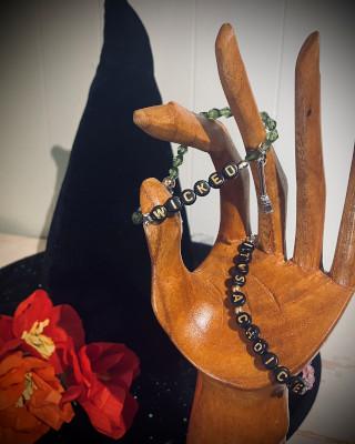 A wooden hand holding two beaded bracelets, one reading Wicked and the other reading It's a Choice. A black witch's hat is in the background.